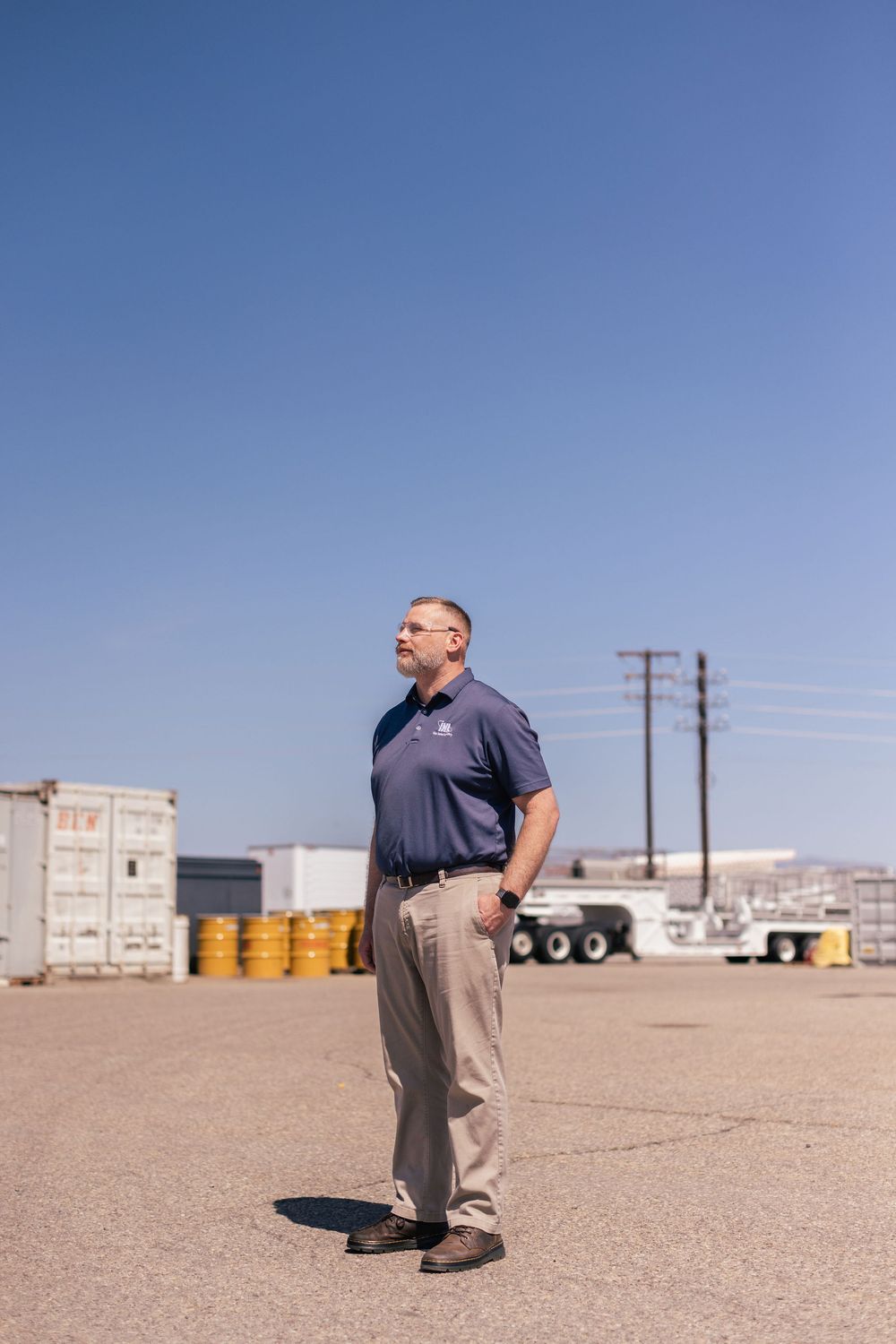 A person stands in a casual stance on an industrial lot with storage containers and utility poles against a bright blue sky.
