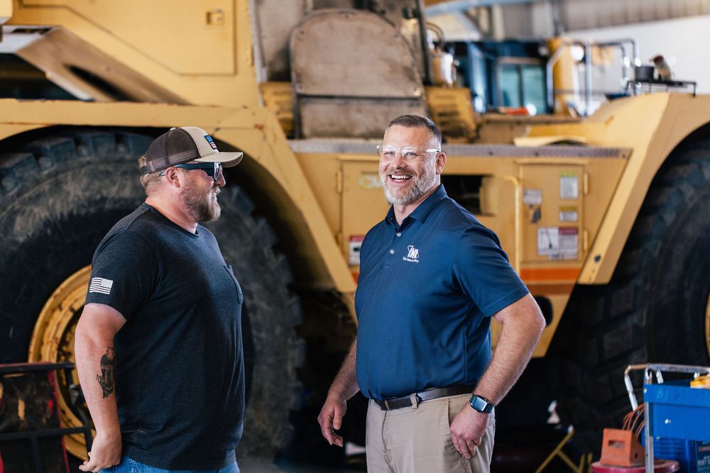 Two construction workers in safety gear discuss plans while standing near heavy yellow equipment.