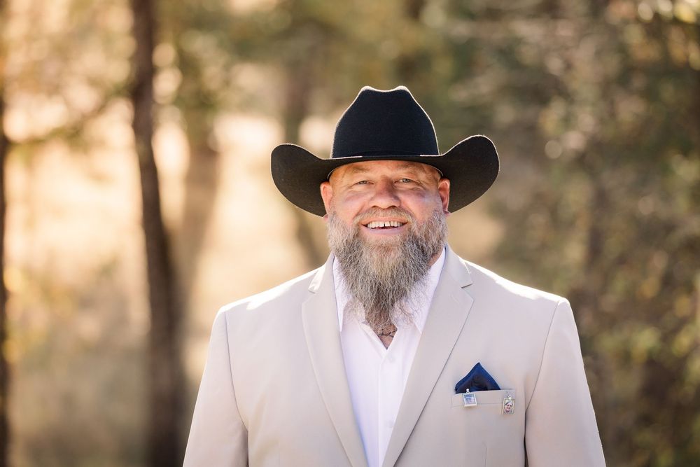 A row of groomsmen in light colored suits and cowboy hats stand outdoors.