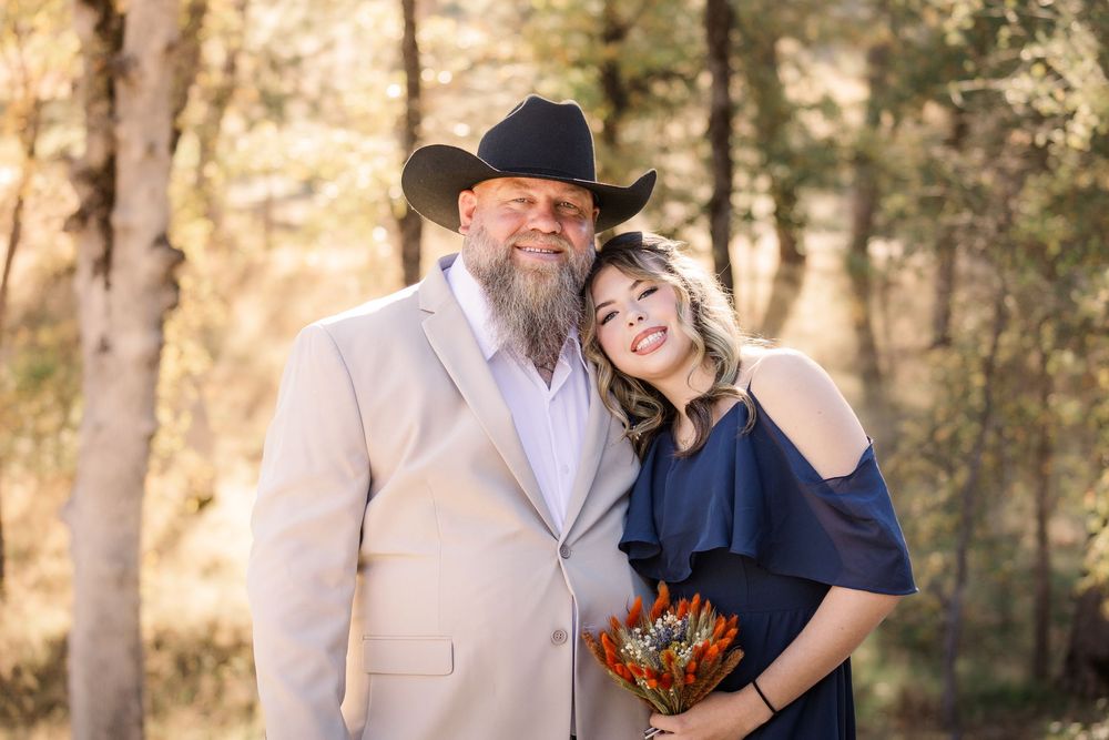 Couple embraces and smiles during outdoor photo session with trees in background.