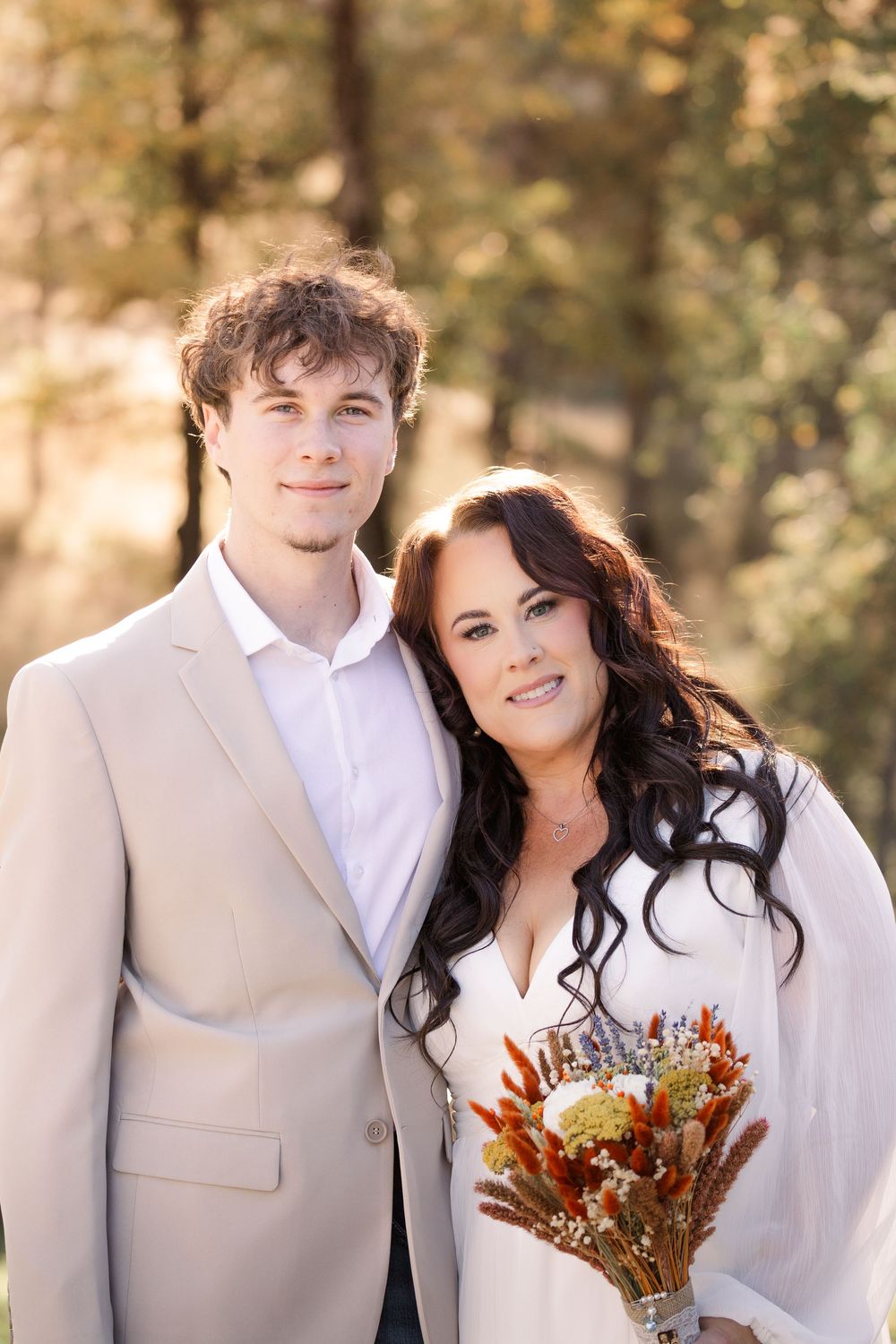 A couple poses together outdoors in white attire with autumn-colored dried flowers during a romantic photoshoot.
