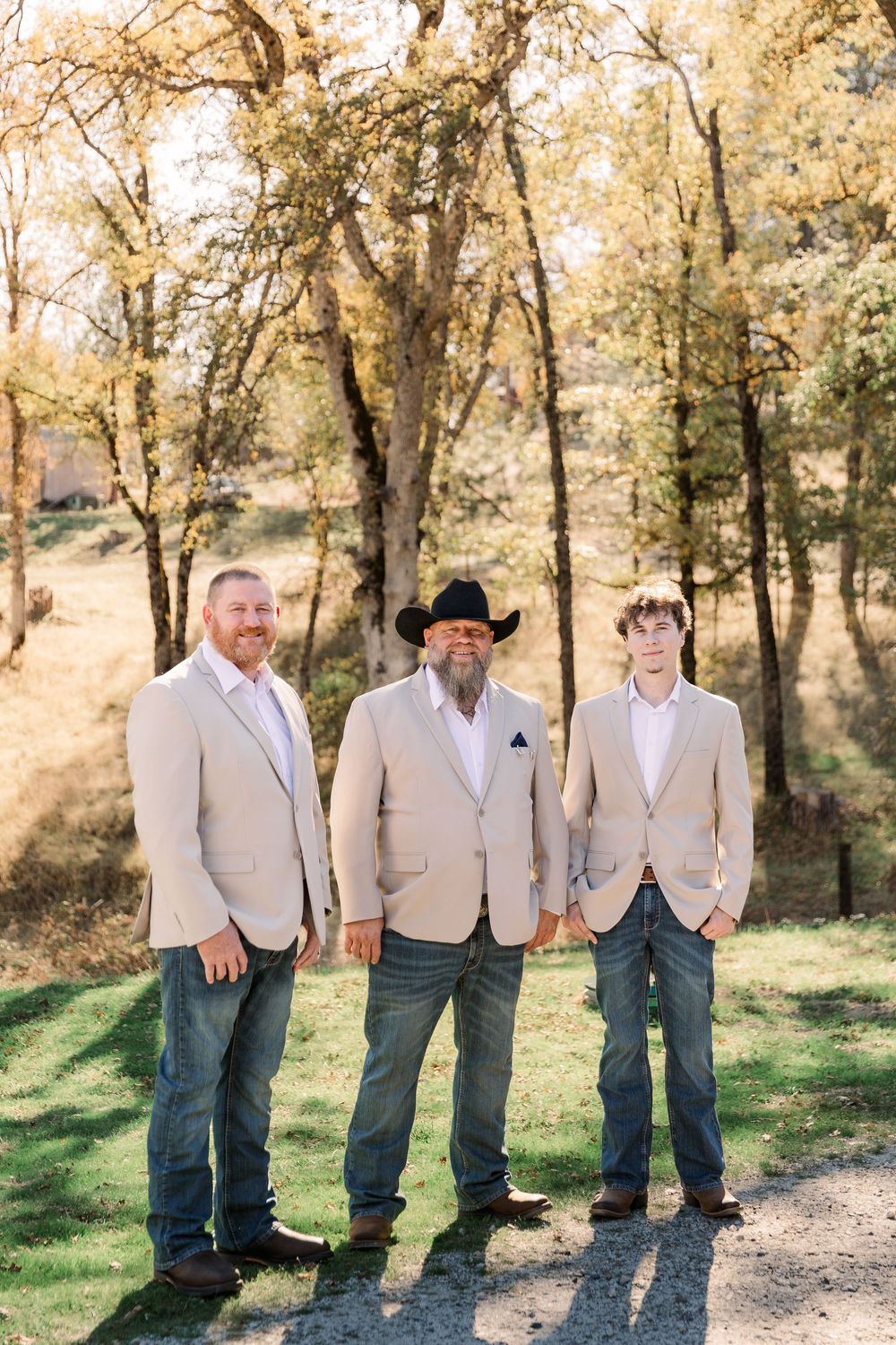 Three groomsmen in beige vests pose together on a dirt path surrounded by autumn trees.