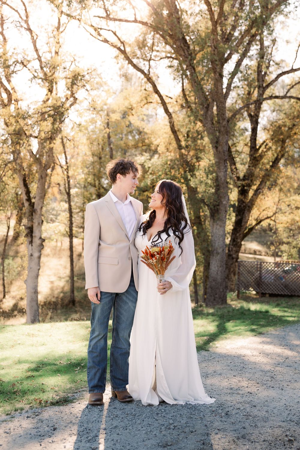A romantic wedding photoshoot on a gravel path surrounded by golden-lit trees in a natural setting.