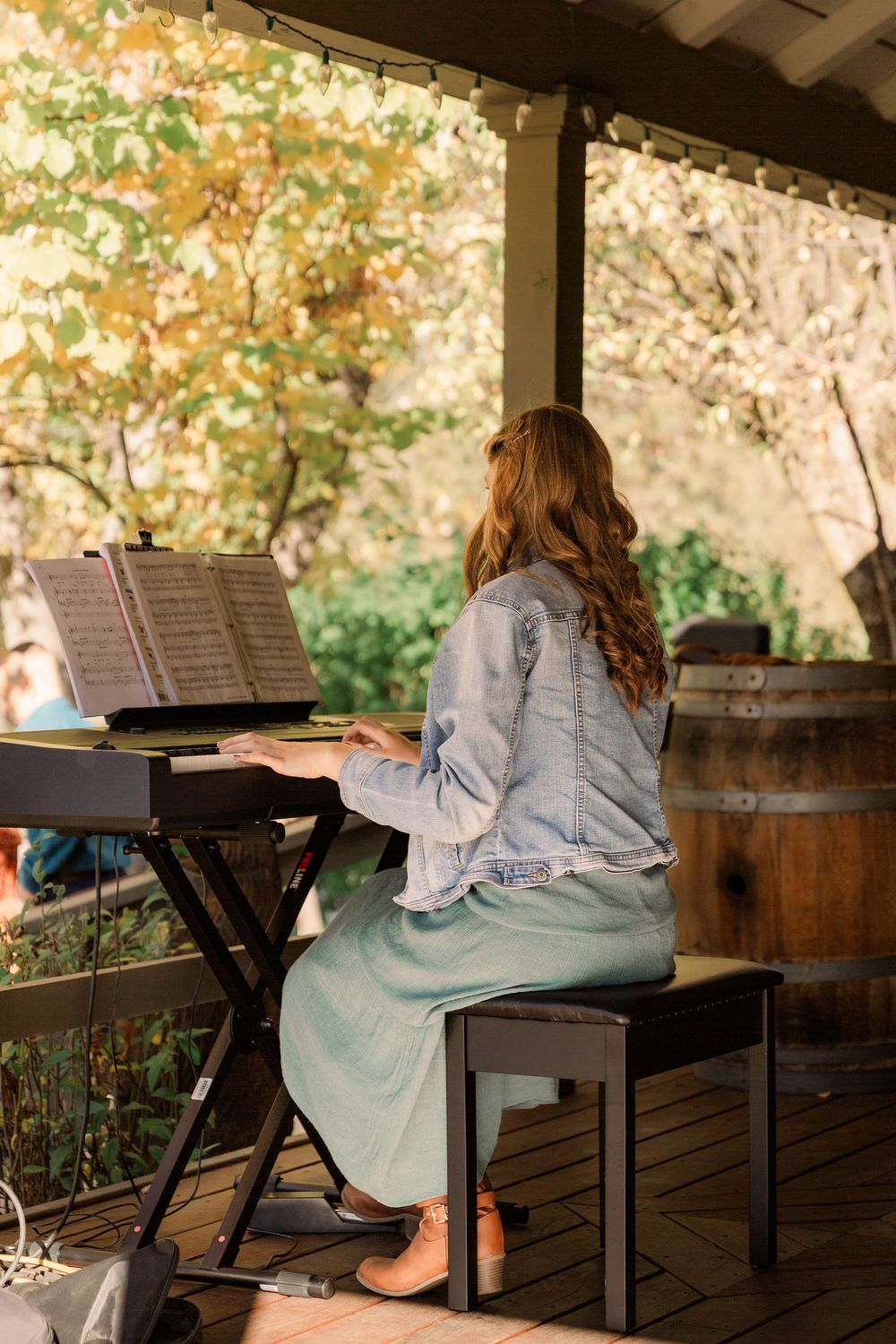 A person sits alone on a wooden bench with a music stand in a covered outdoor setting during autumn.