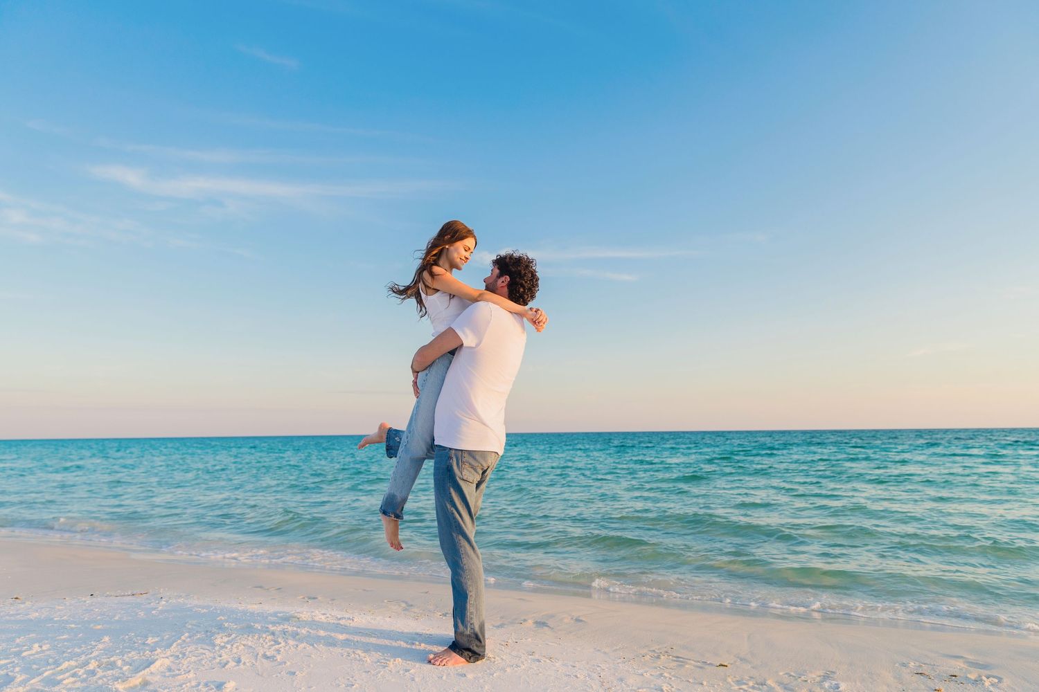 A couple embraces playfully on a pristine beach with turquoise waters and clear blue sky in the background.