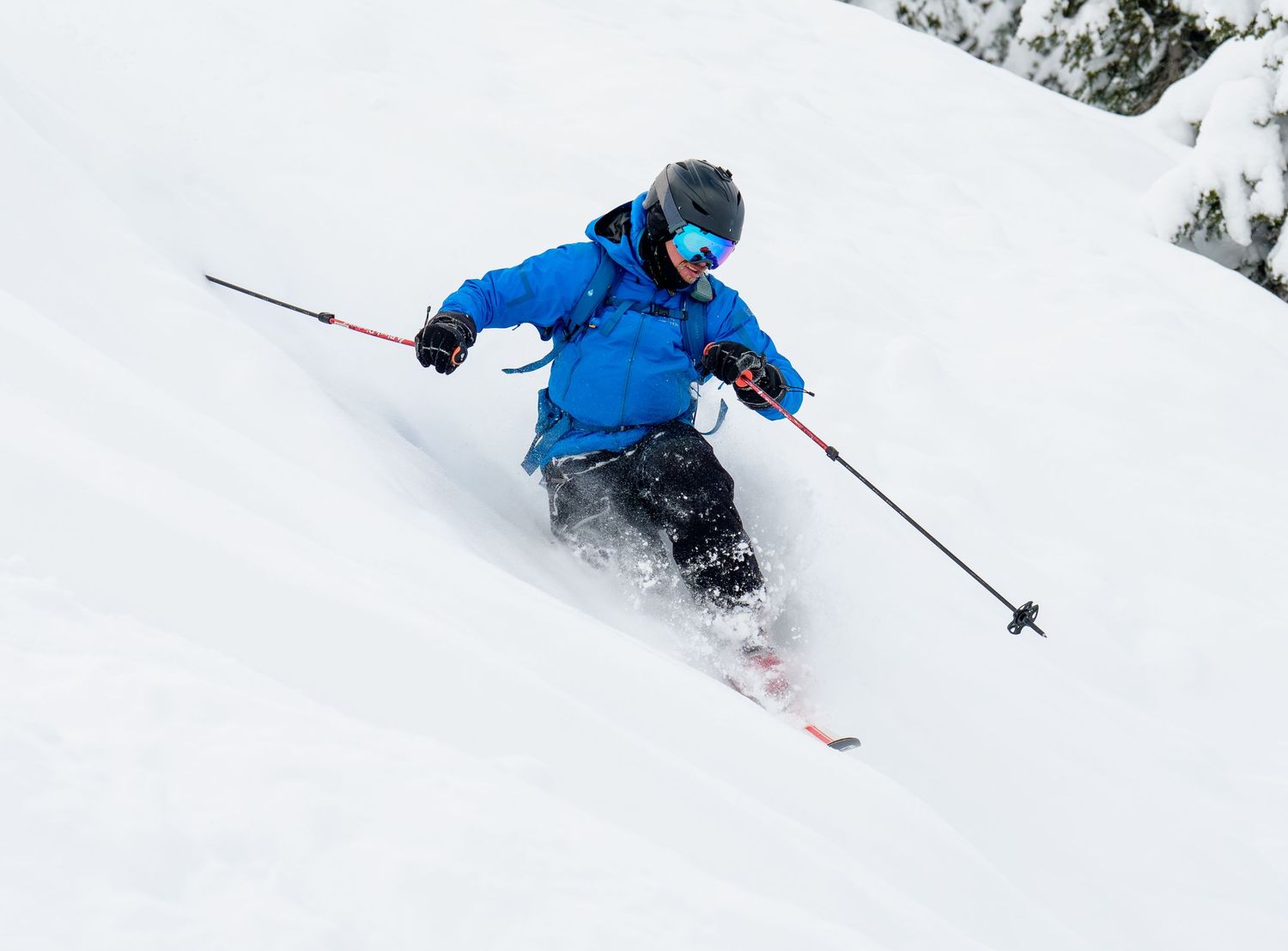 A skier carves through fresh powder snow on a steep mountain slope in a bright blue jacket.