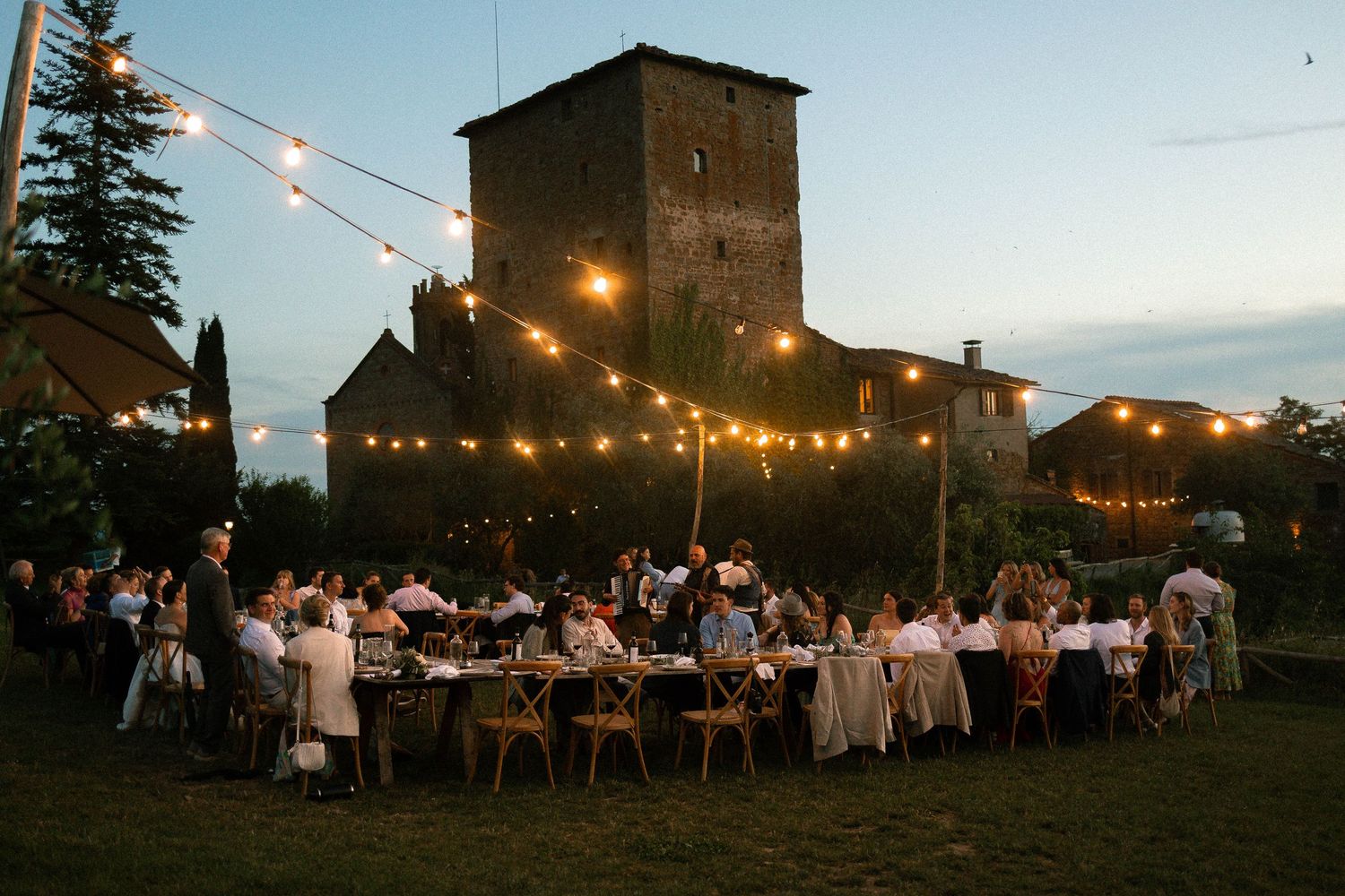 String lights illuminate an outdoor evening wedding reception with long tables set up on a lawn near a stone tower.