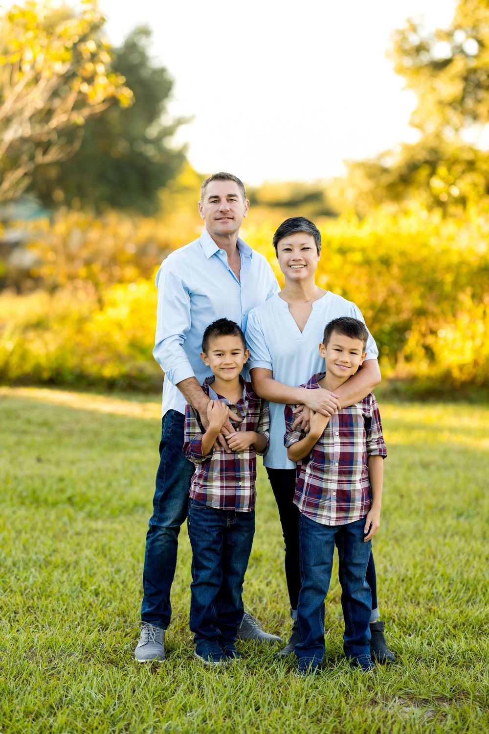 Family wearing coordinated plaid shirts and jeans poses together on grassy hill during golden hour sunset.
