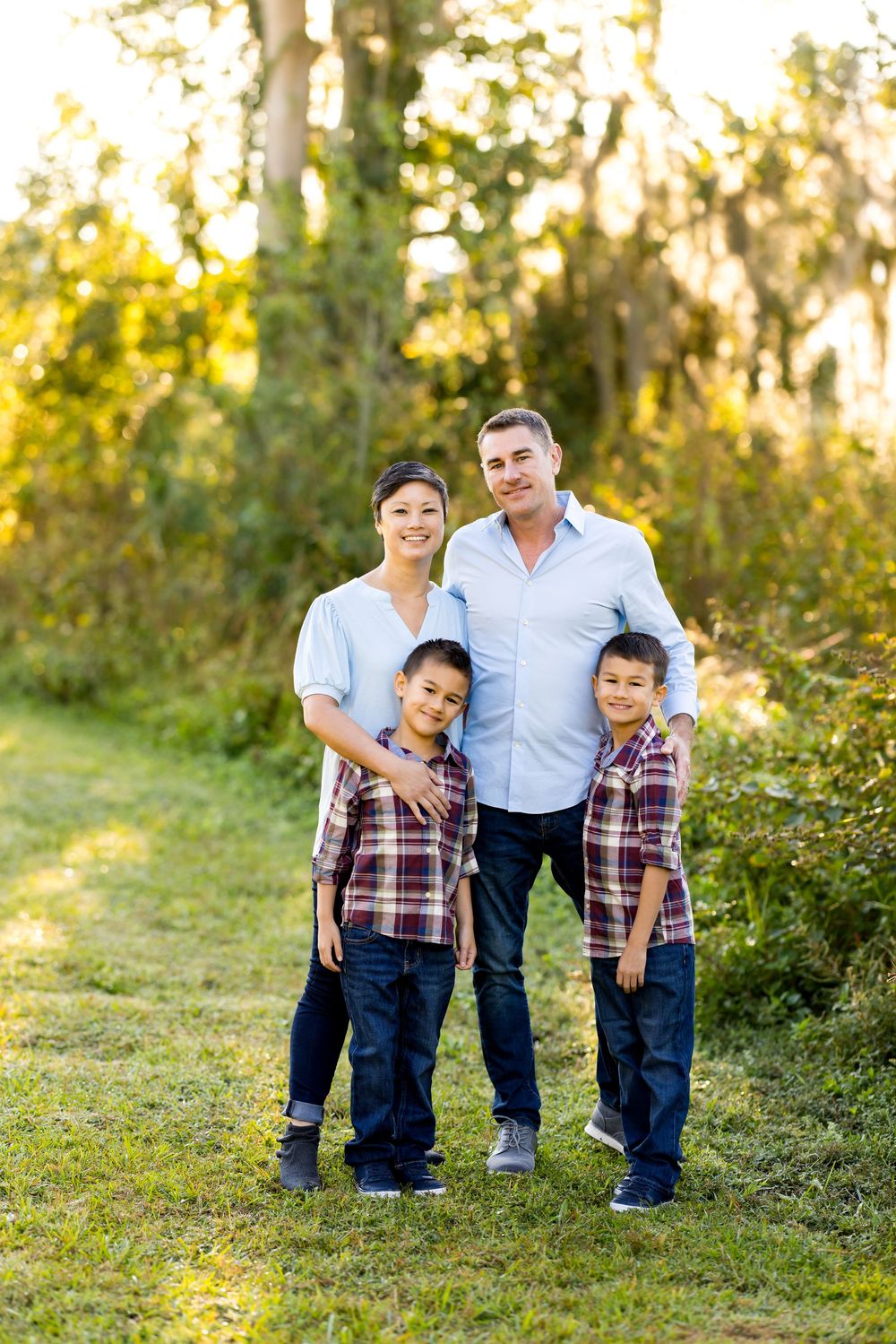 Family of four wearing coordinated blue and plaid outfits poses together outdoors in golden sunset lighting.