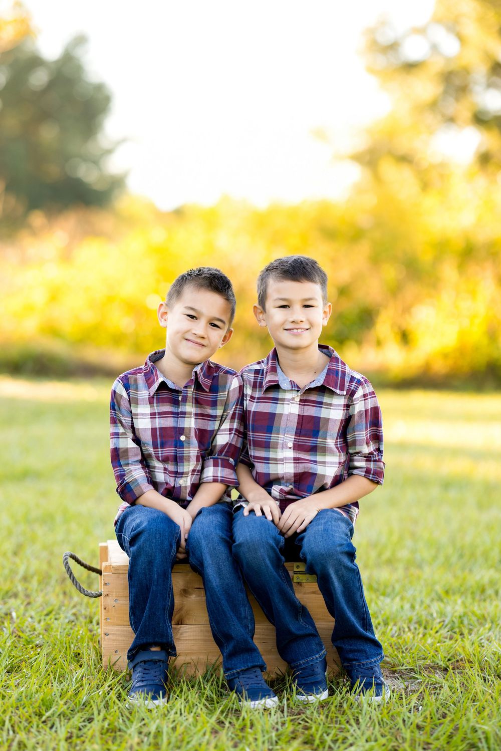 Two children in matching plaid shirts sit together on a wooden bench in a sunlit grassy field.