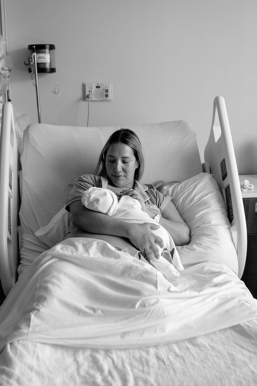 A patient rests peacefully in a hospital bed holding a newborn baby wrapped in white blankets.