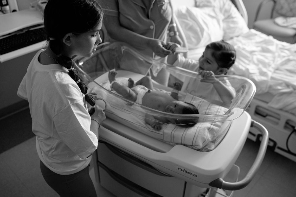 Medical professionals in white coats examine newborn babies in hospital bassinets during routine care.