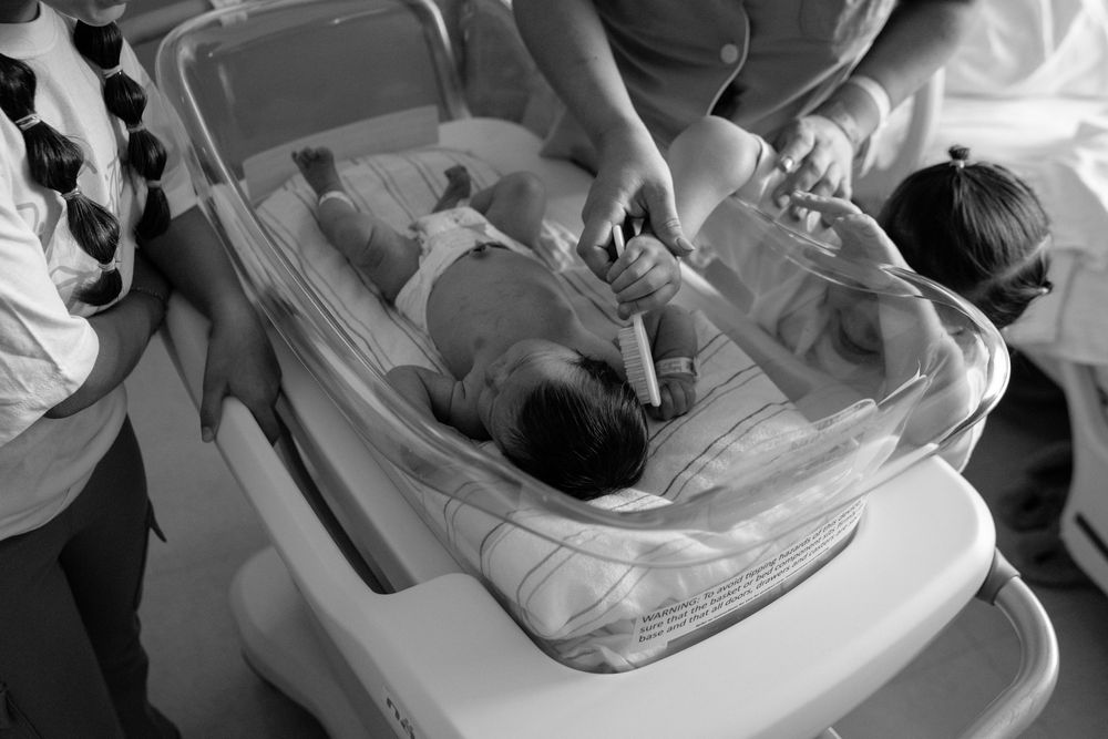 A newborn baby lies in a clear hospital bassinet while being examined.