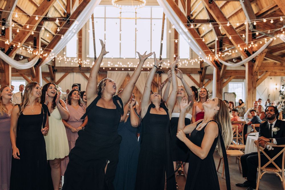 Women in formal dresses reaching up to catch a wedding bouquet toss in a rustic barn venue with string lights.