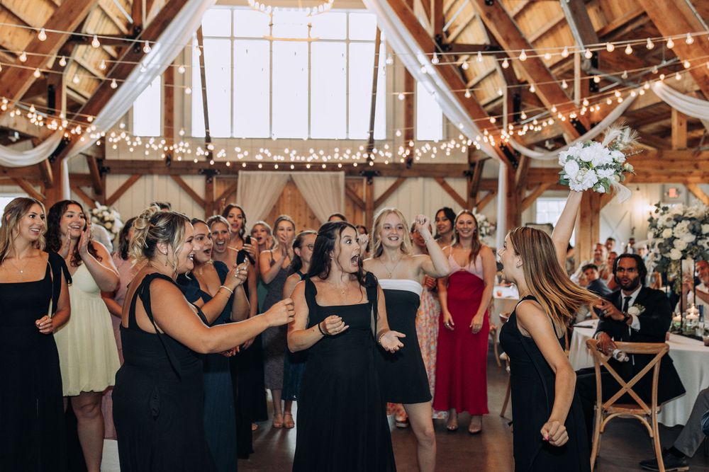 Excited wedding guests in elegant dresses react to a bouquet toss inside a decorated barn reception hall.