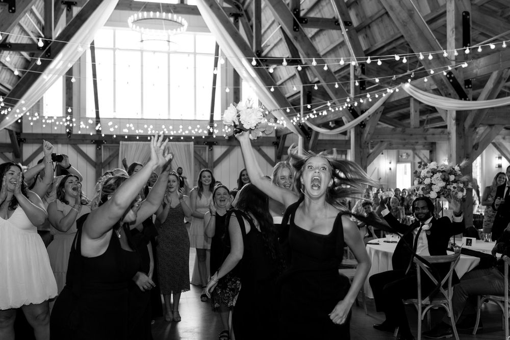 Black and white photo of joyful women competing to catch a bridal bouquet at a barn wedding reception.