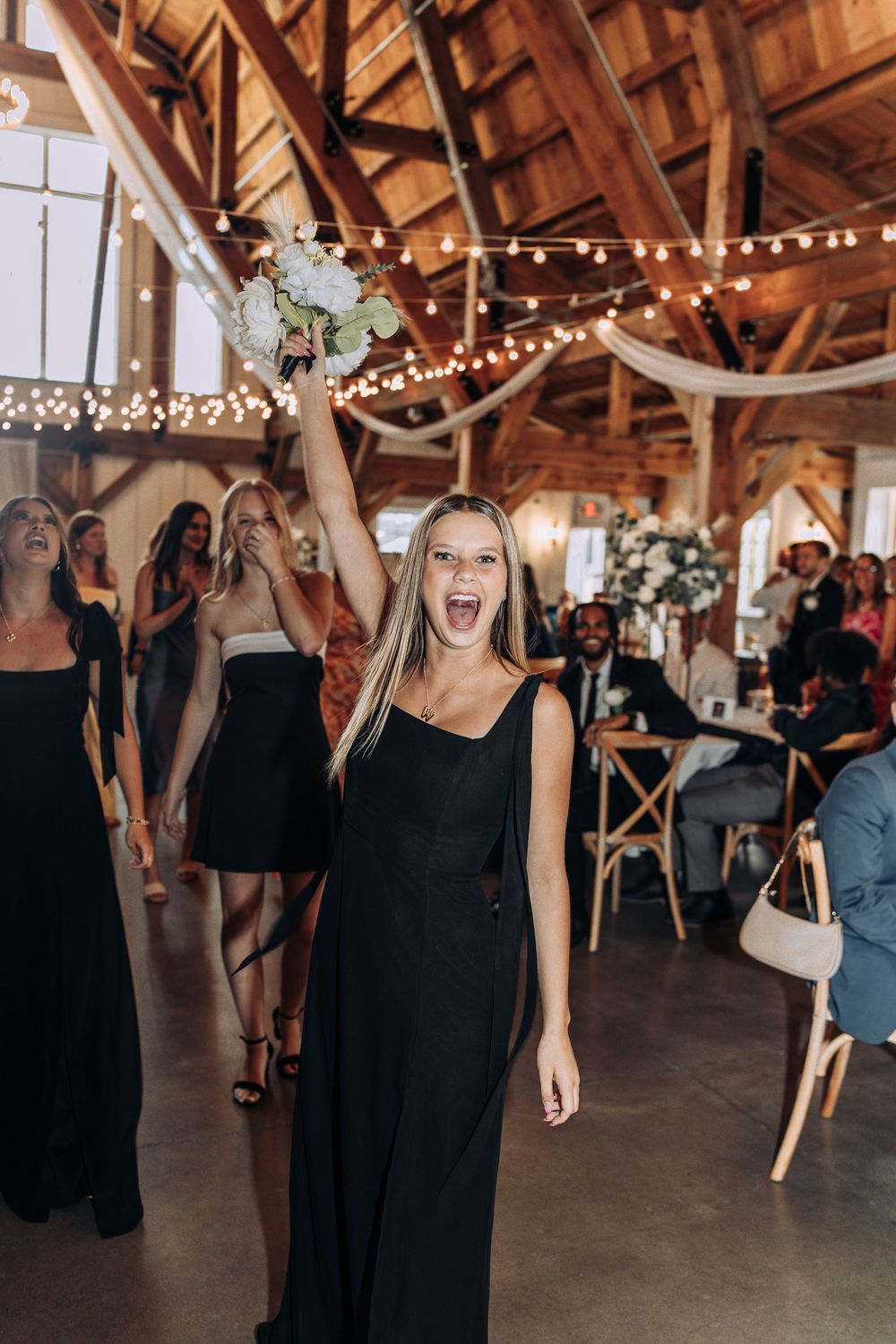 Excited woman in black dress triumphantly holds caught wedding bouquet aloft in a rustic barn venue.