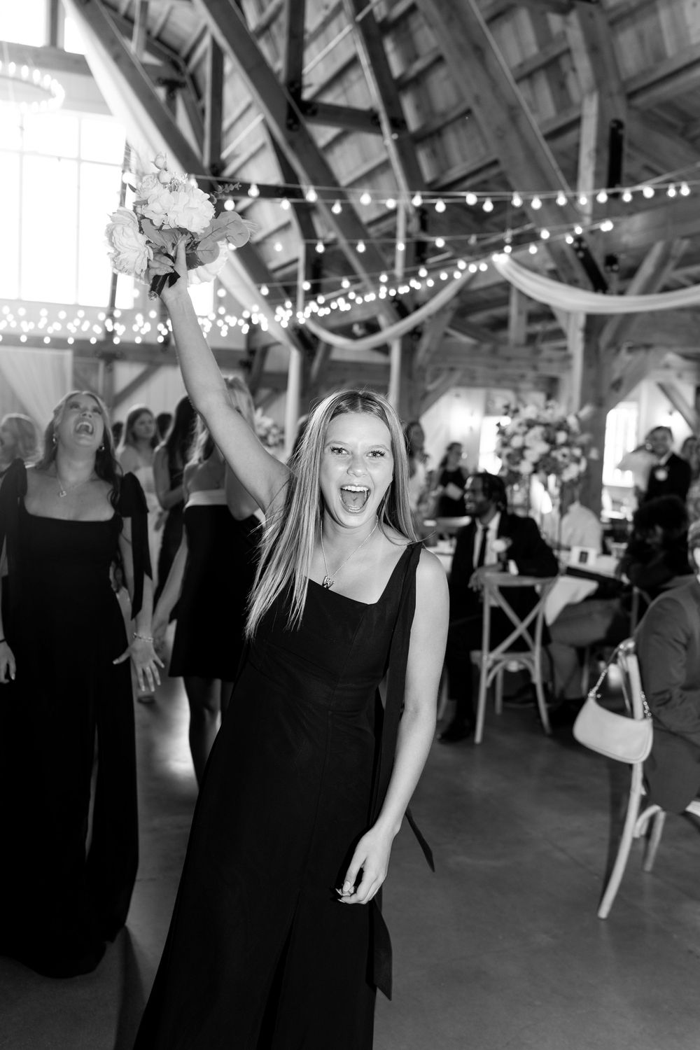 Black and white image of smiling woman raising caught bridal bouquet in celebration at barn wedding.