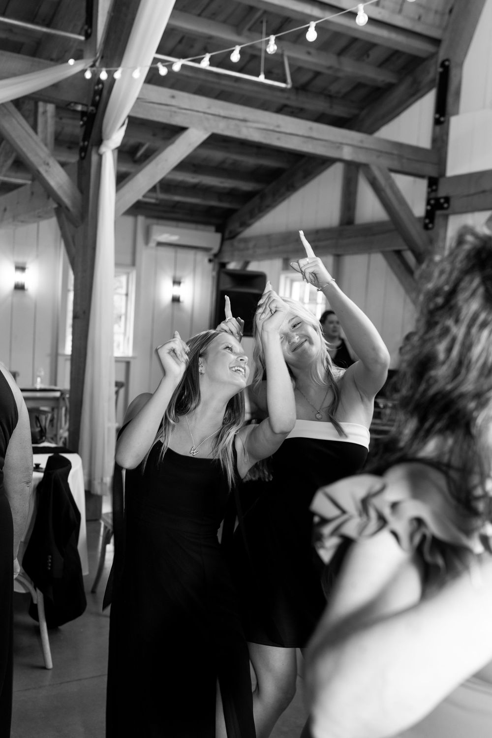Two laughing women in black dresses point upward together at a rustic barn wedding reception.