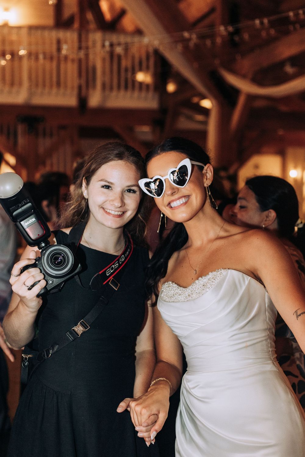 Wedding photographer poses with smiling bride wearing heart-shaped sunglasses at a rustic barn reception.