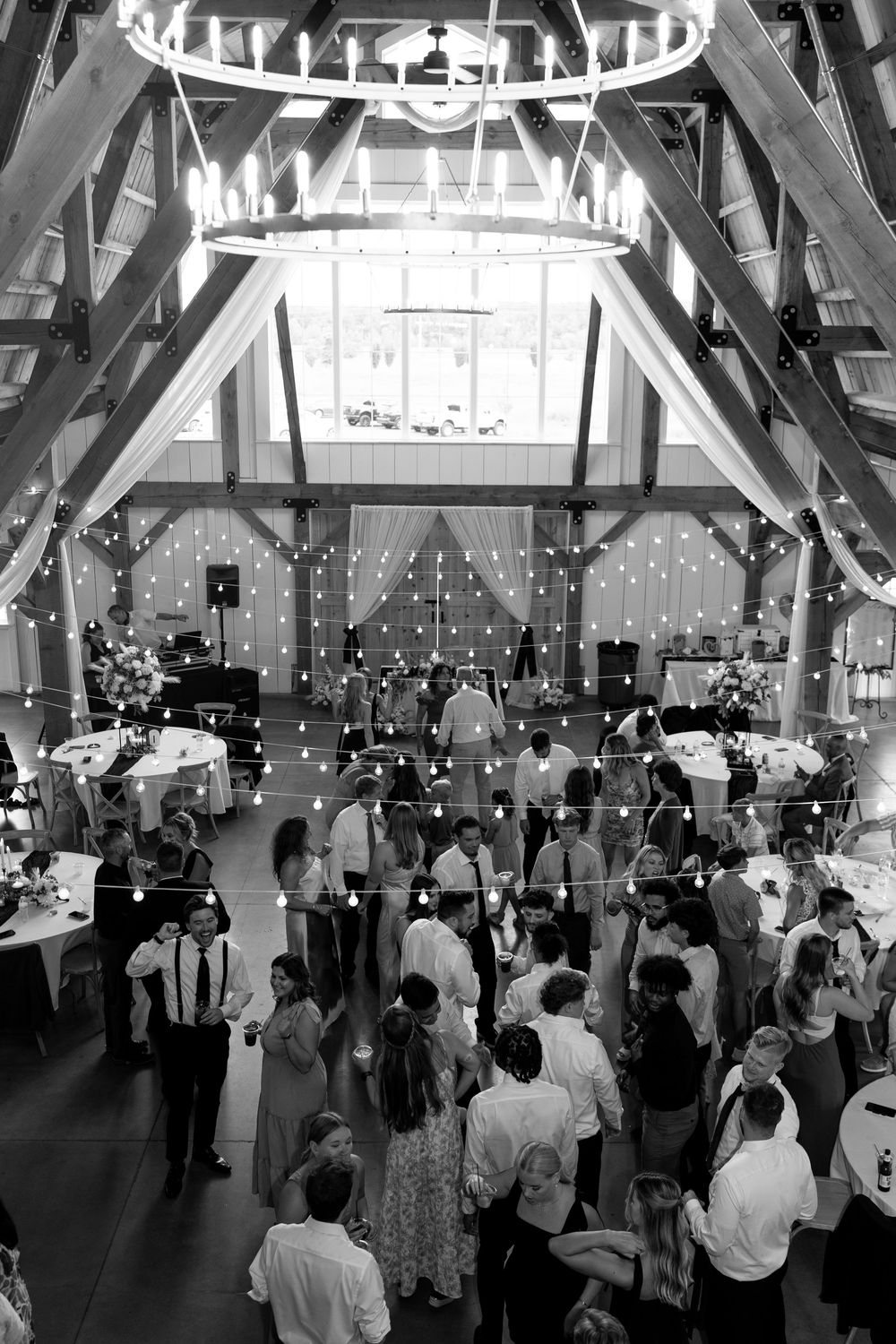 Black and white aerial view of wedding guests dancing under string lights in an elegant barn venue.