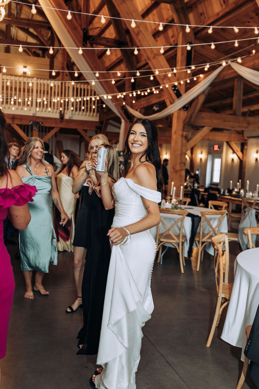 Elegant bride in white off-shoulder gown smiles while walking through her rustic barn wedding reception.