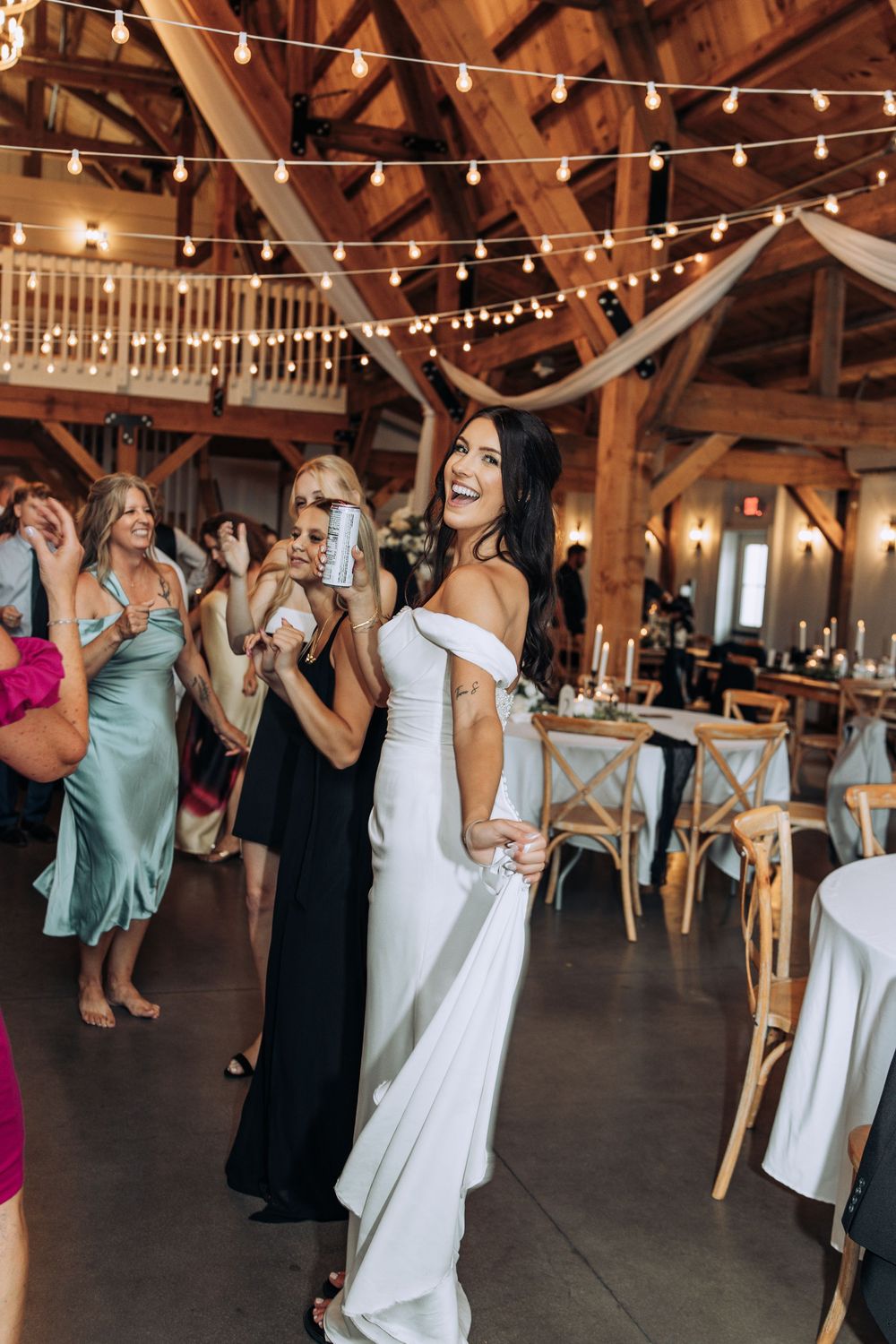 Bride in white off-shoulder gown laughing and dancing at rustic barn wedding reception with string lights.