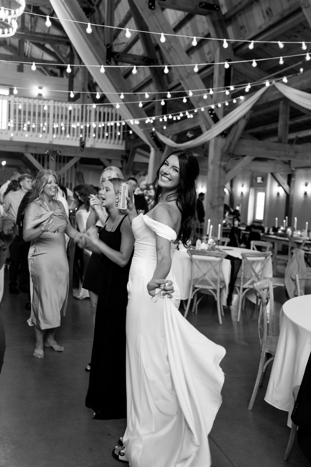Black and white photo of bride in elegant white gown dancing joyfully at rustic barn wedding reception.