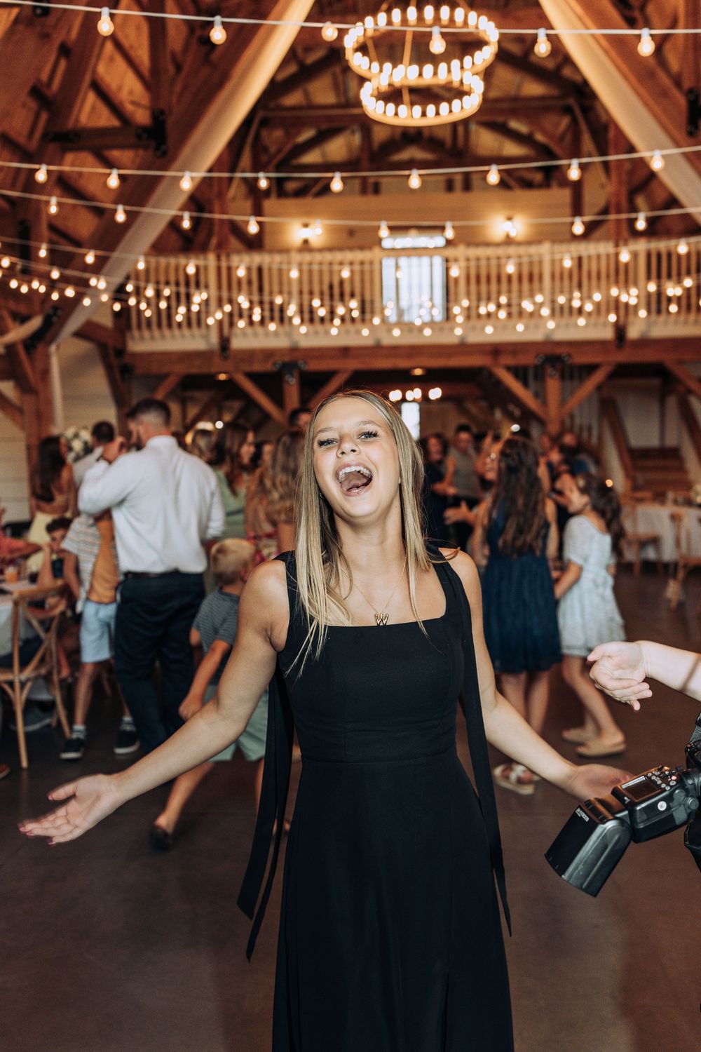 Excited blonde guest in black dress singing and dancing at lively rustic barn wedding reception.