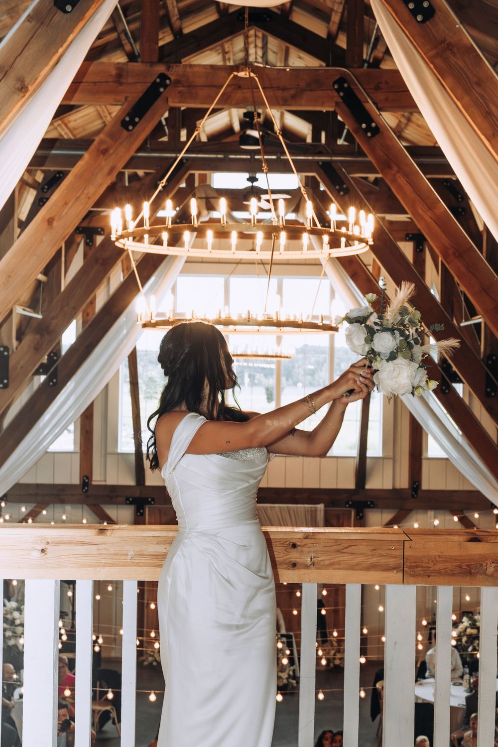 Bride holds white floral bouquet beneath rustic wooden barn ceiling with candle chandelier and lights.