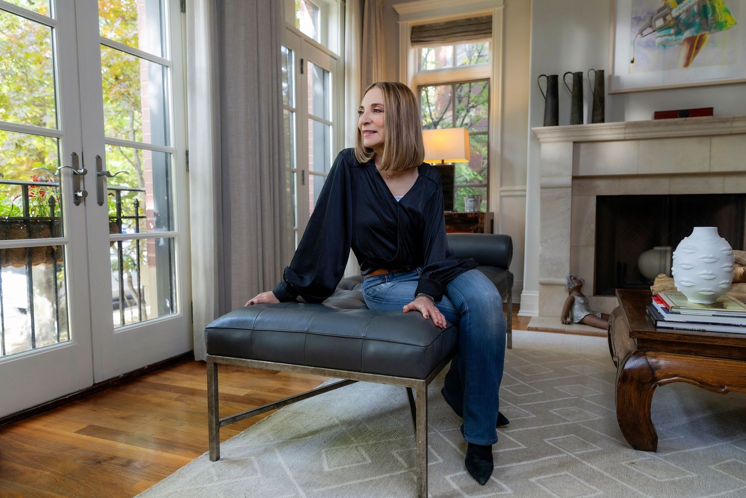 A person in a navy blazer and white top relaxes on a brown leather couch in a bright, airy living room.