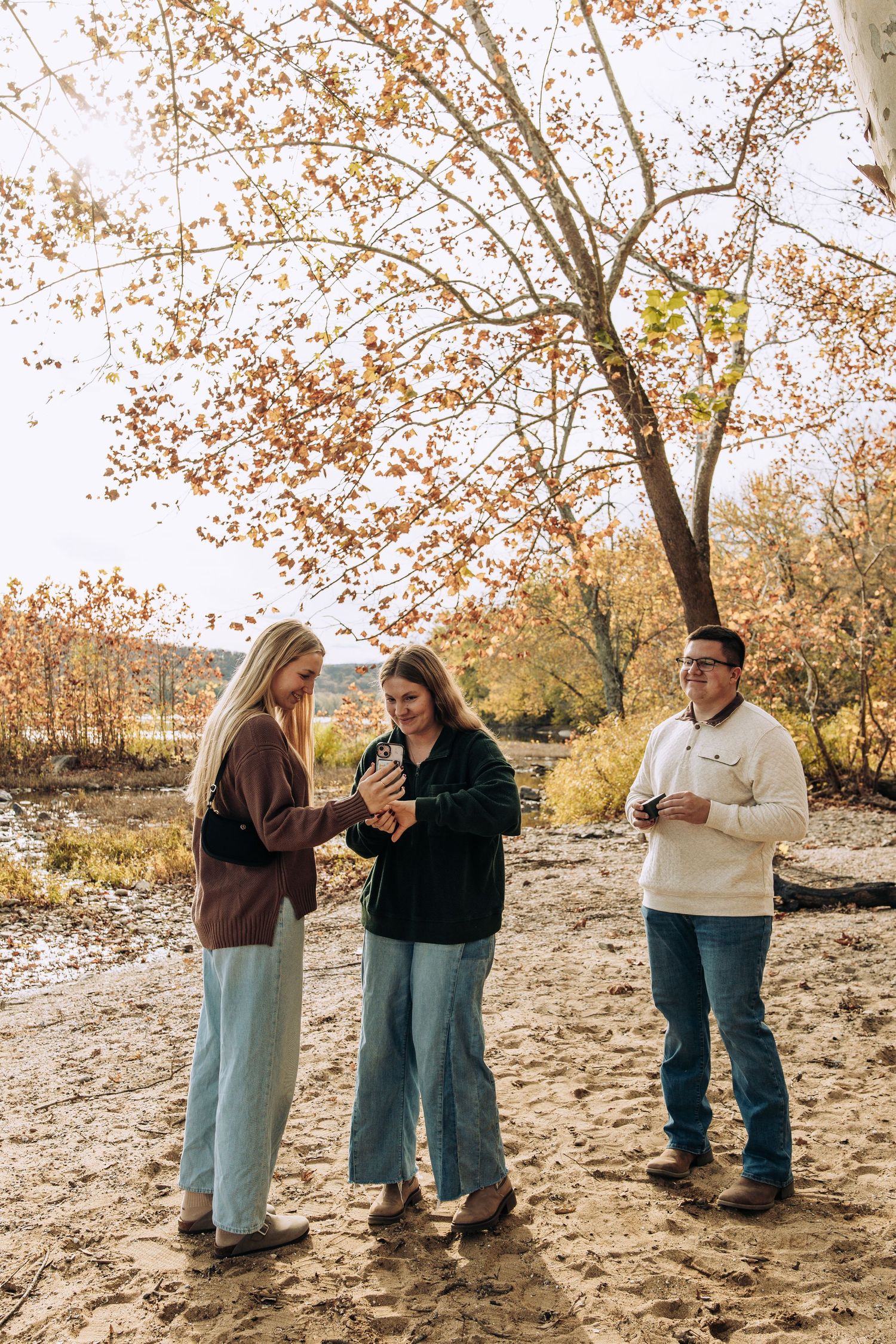 Three friends gather on a sandy riverbank examining a camera amid colorful fall foliage.