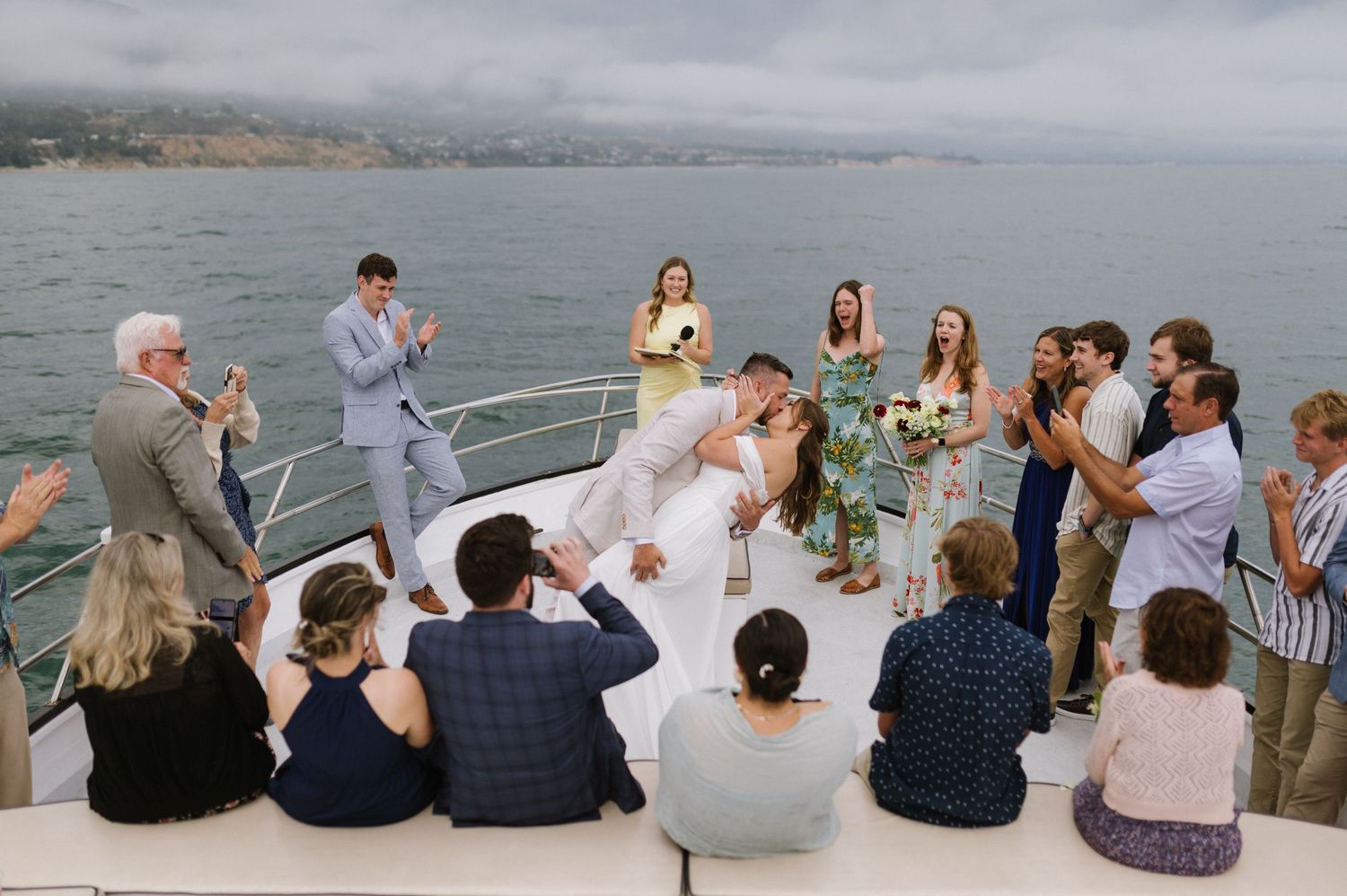 Wedding guests gather on a boat deck overlooking a scenic coastal bay during a maritime celebration ceremony.