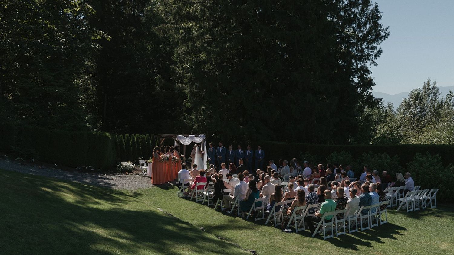 Wide aerial view of elegant outdoor wedding ceremony with guests seated on white chairs among tall trees.