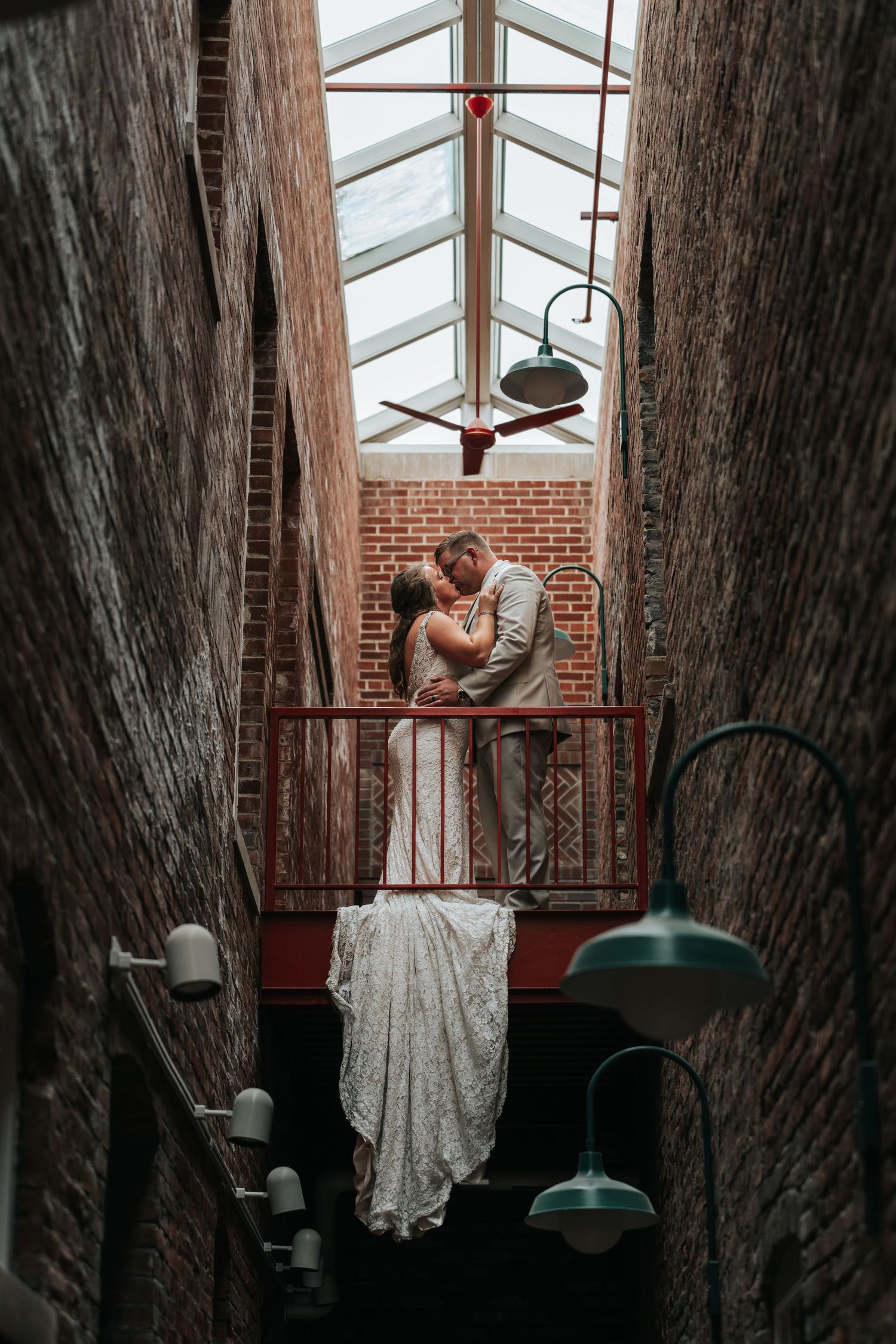 Wedding couple embracing in a brick-walled sunroom with industrial lighting fixtures.