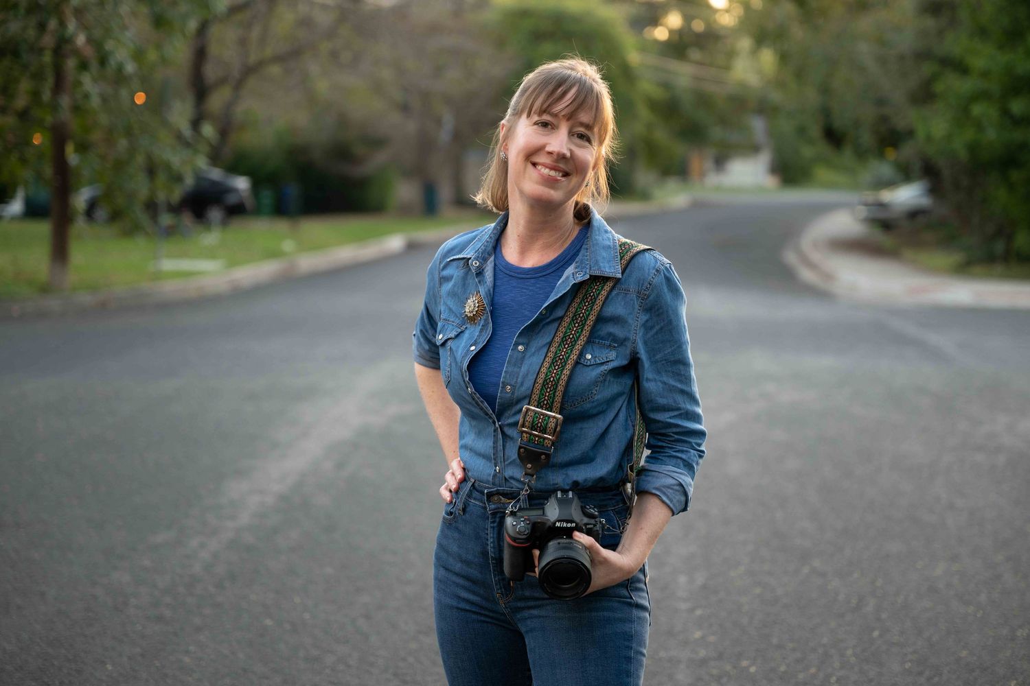 A photographer wearing denim stands on a quiet suburban street with a camera and strap.