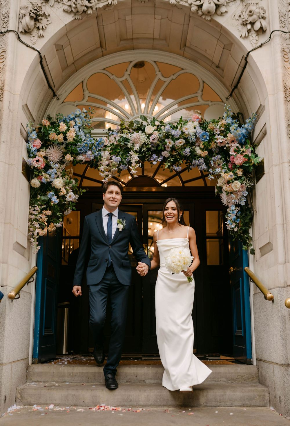 Wedding couple poses under ornate stone archway decorated with floral arrangements and greenery.