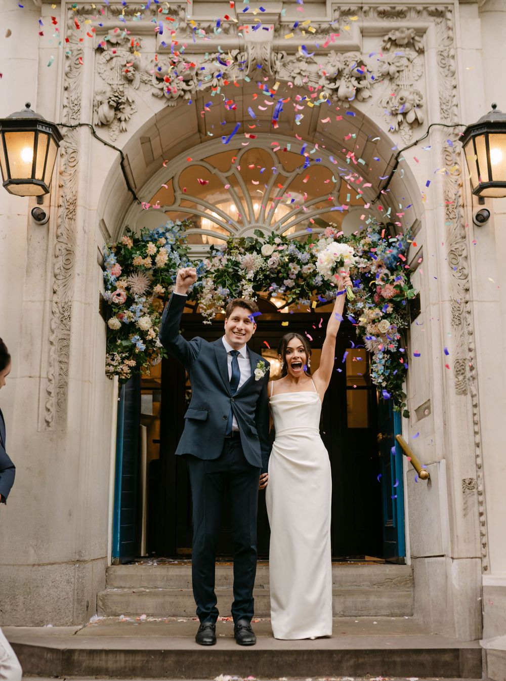 Confetti falls as newlyweds celebrate under decorated stone archway with floral arrangements.