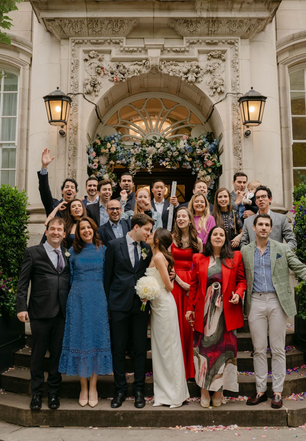 Wedding group portrait on stone steps with guests in colorful attire under ornate architecture.
