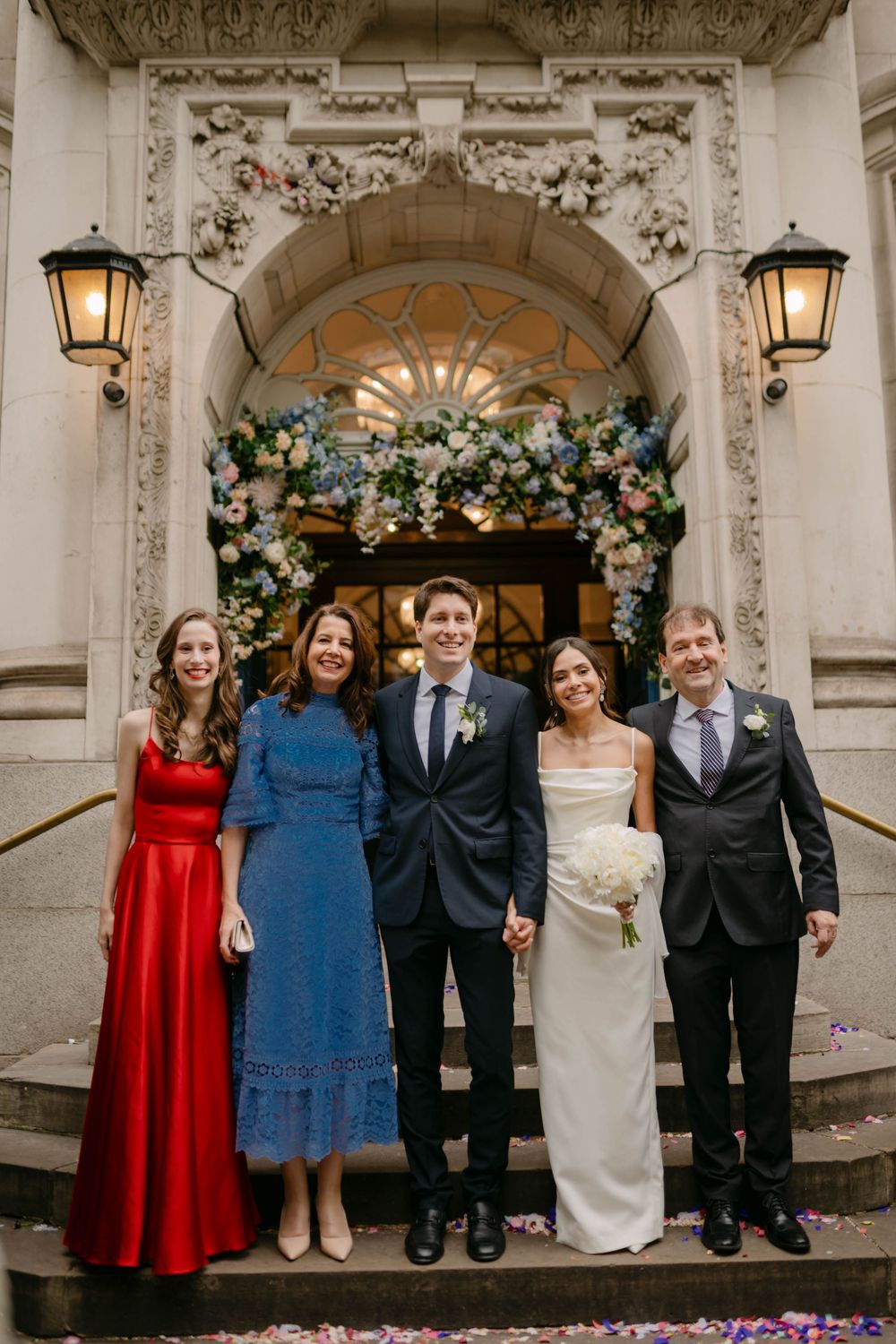 A wedding party poses on stone steps under an ornate floral arch with lanterns.