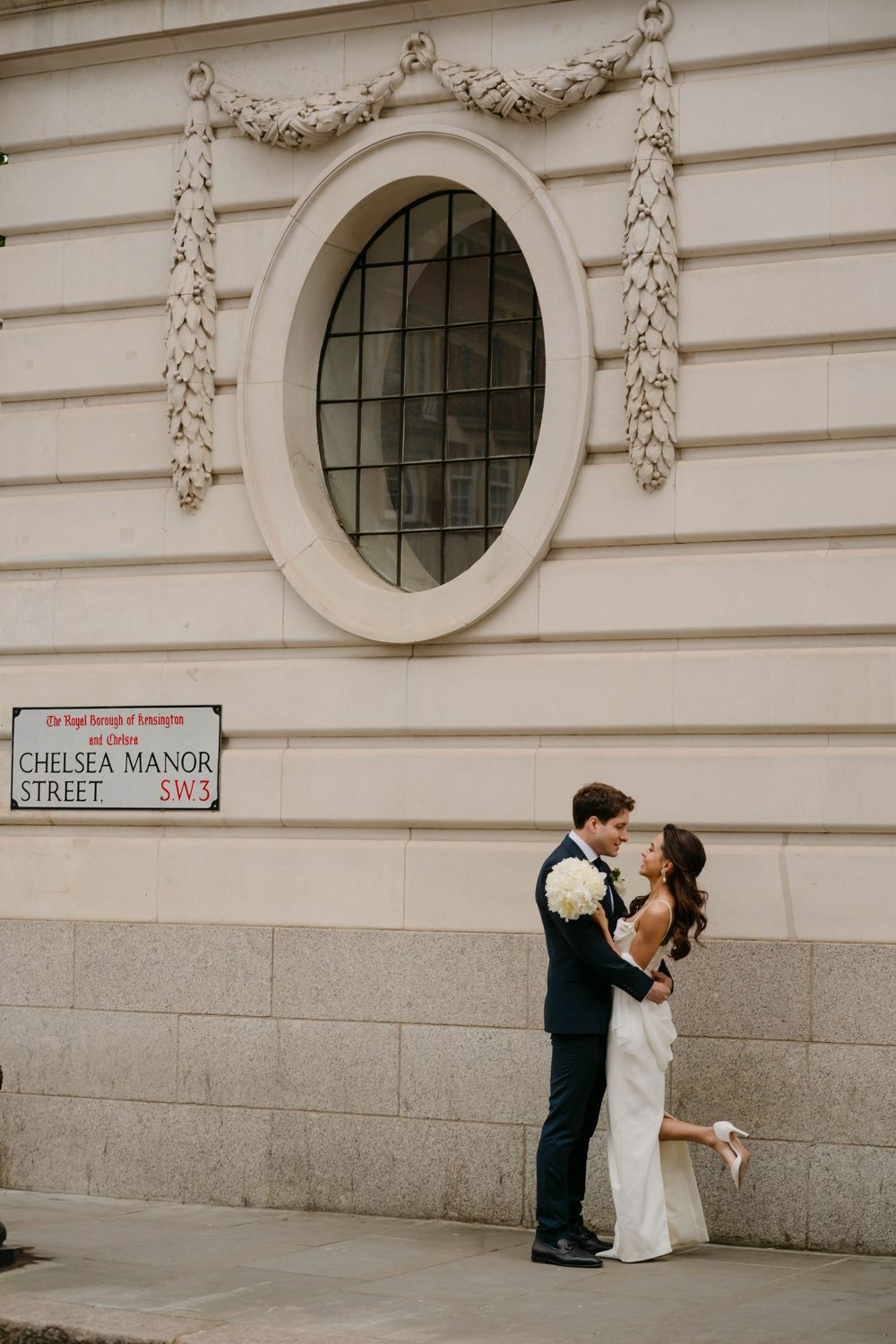 A couple shares a romantic moment against an ornate white building facade with a Chelsea Manor Street sign.