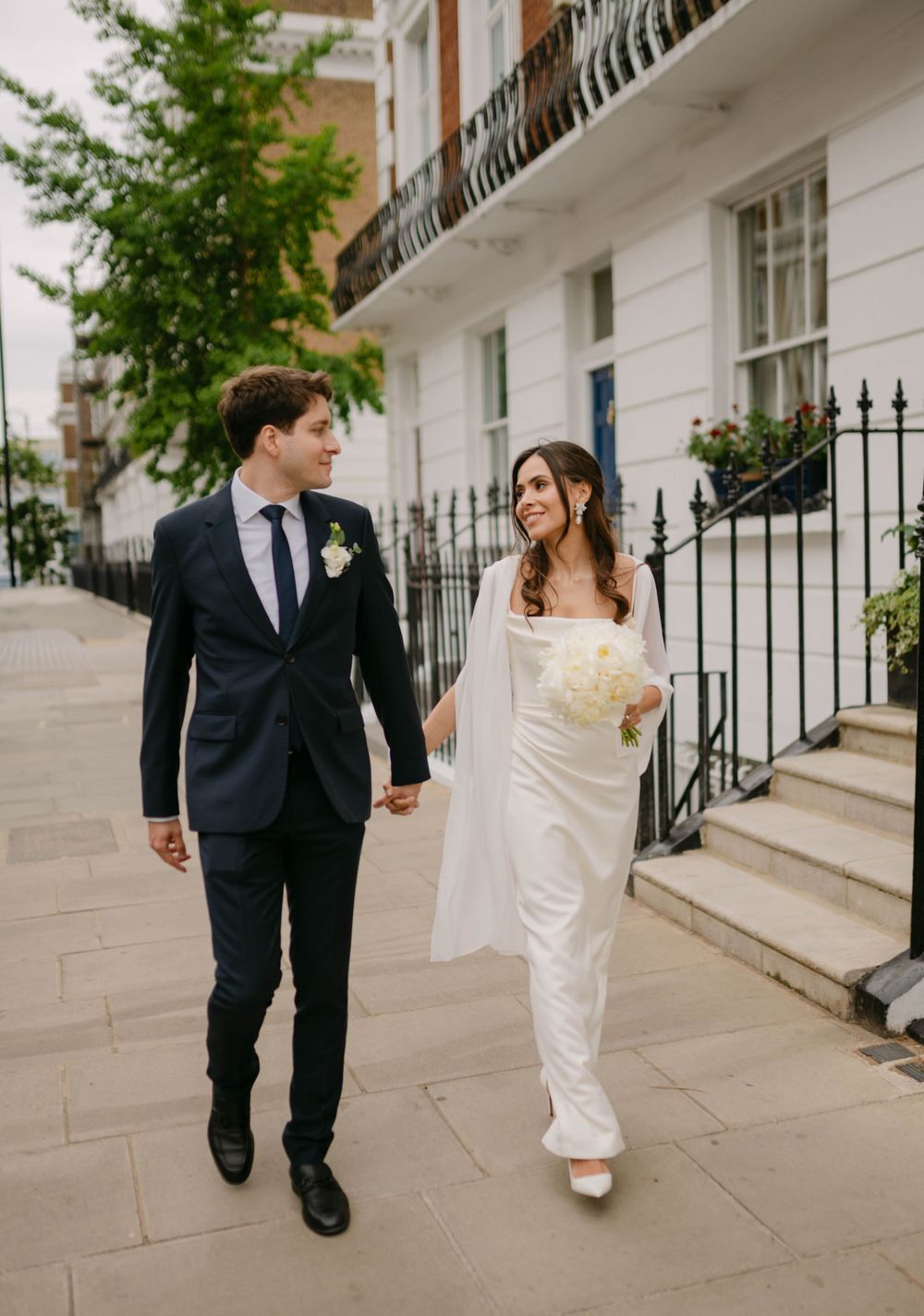 A newlywed couple walks playfully along a London sidewalk lined with white townhouses.