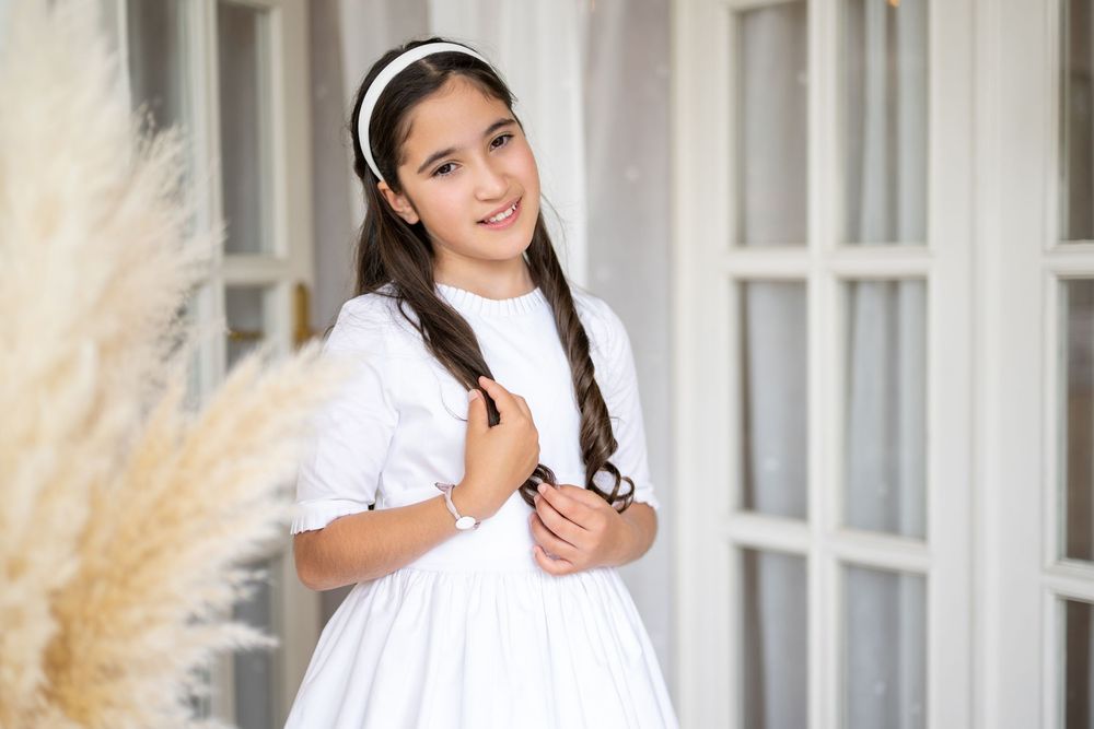 Woman in white dress poses outdoors against light-colored wall with decorative pampas grass.