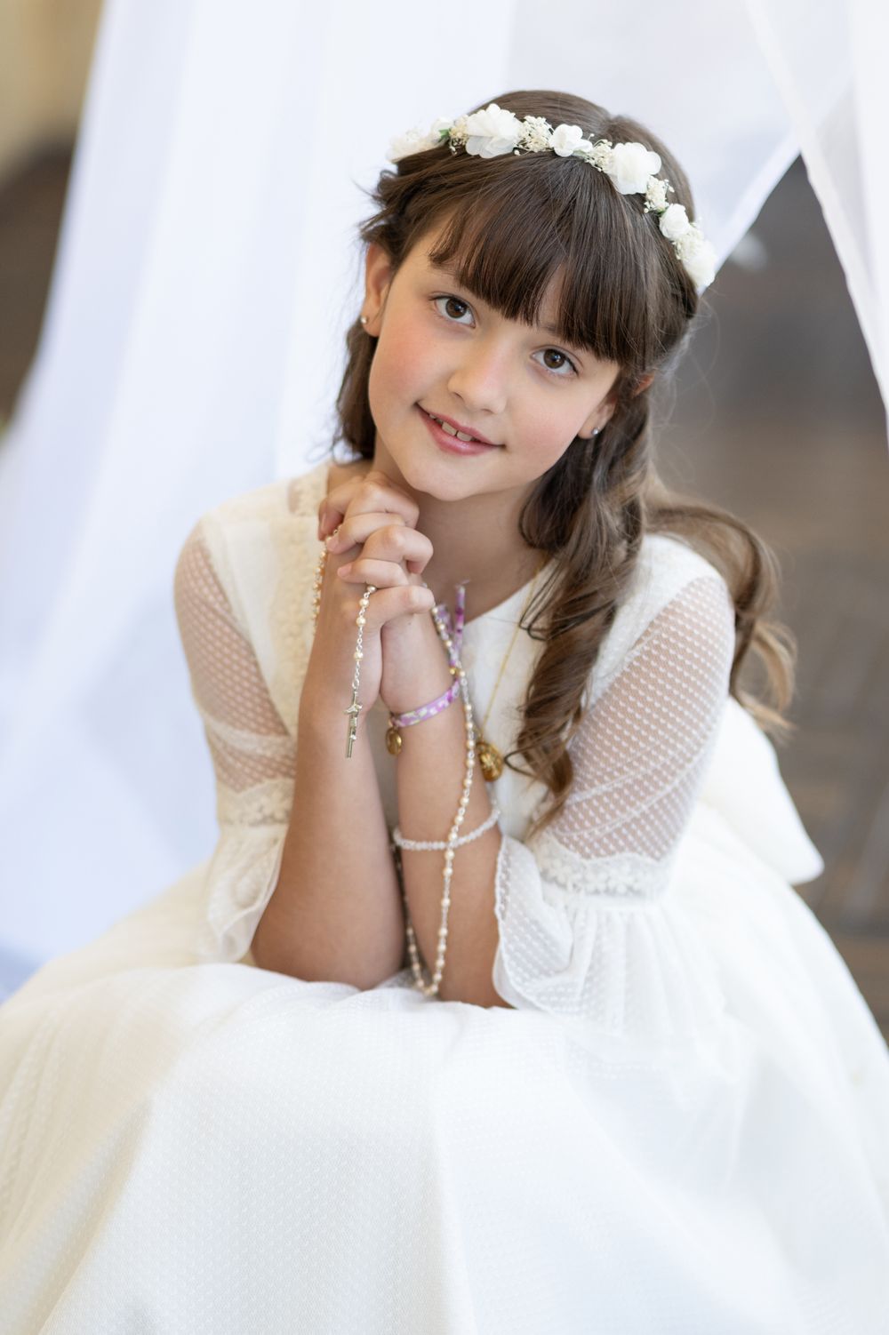 Young girl in white First Communion dress and floral crown poses elegantly with rosary beads.