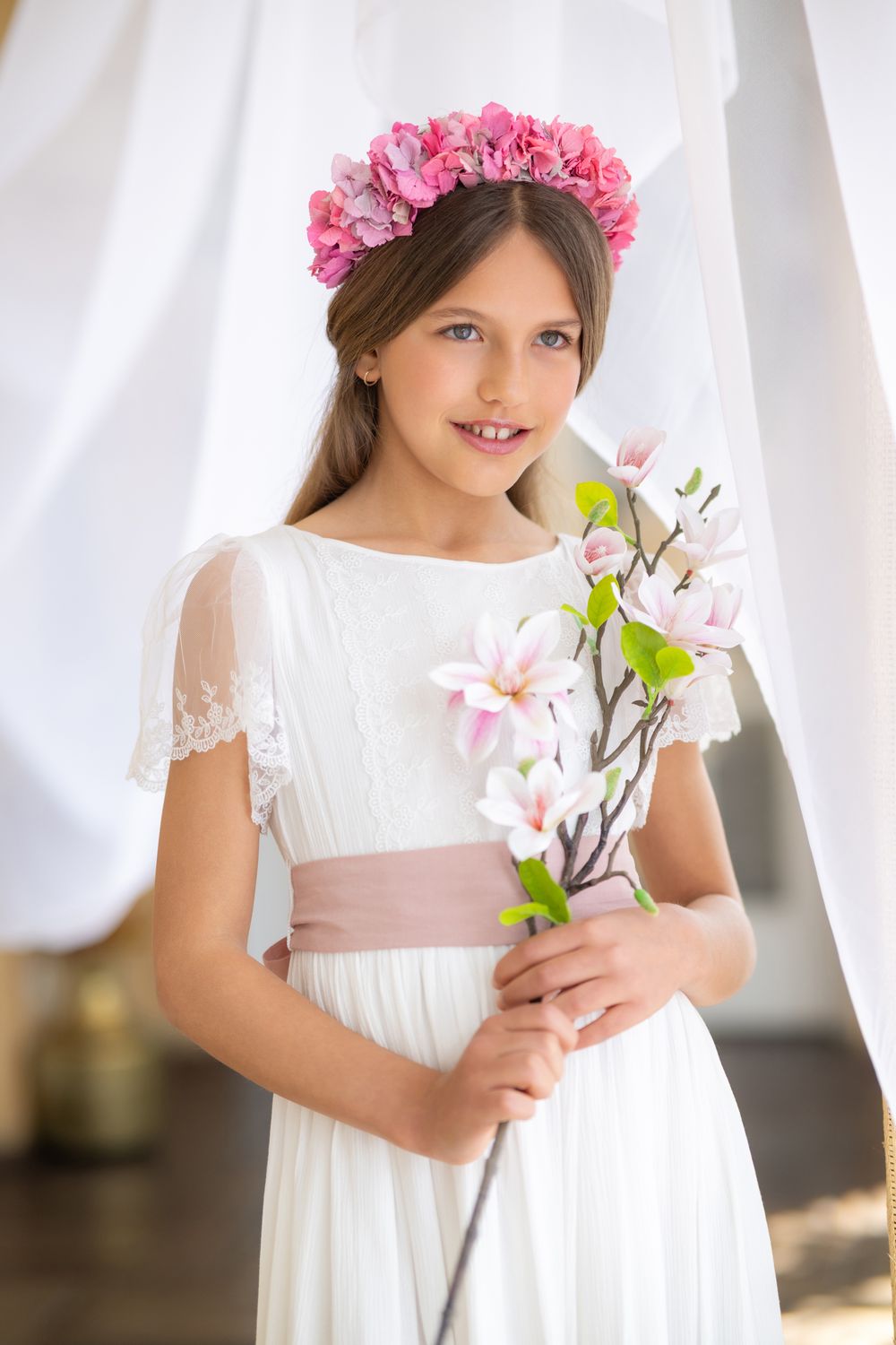 A child in a white dress with a pink sash holds spring flowers while wearing a pink floral crown.