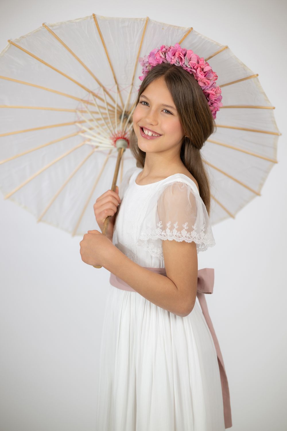 Young girl in white dress poses with paper parasol and pink floral crown in studio photo series.