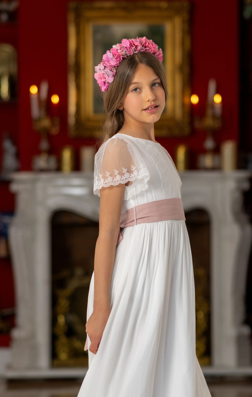 Young girl in a flowing white empire waist dress with pink floral crown and sash poses in an elegant setting.