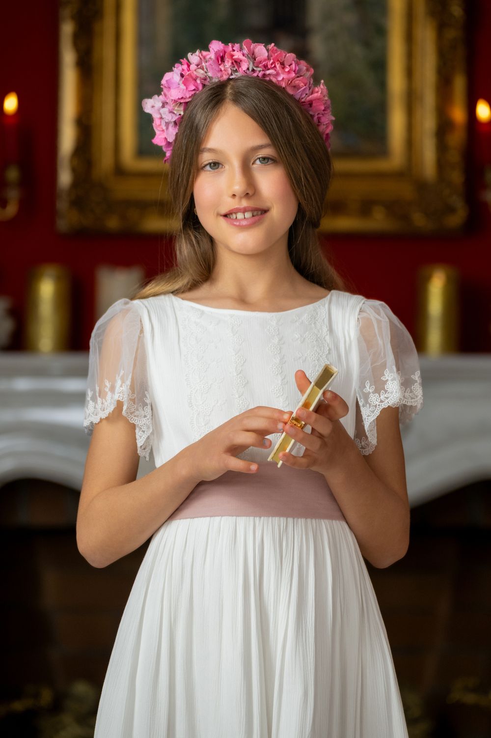 A young person in a white lace dress and pink flower crown holds something delicately in their hands.