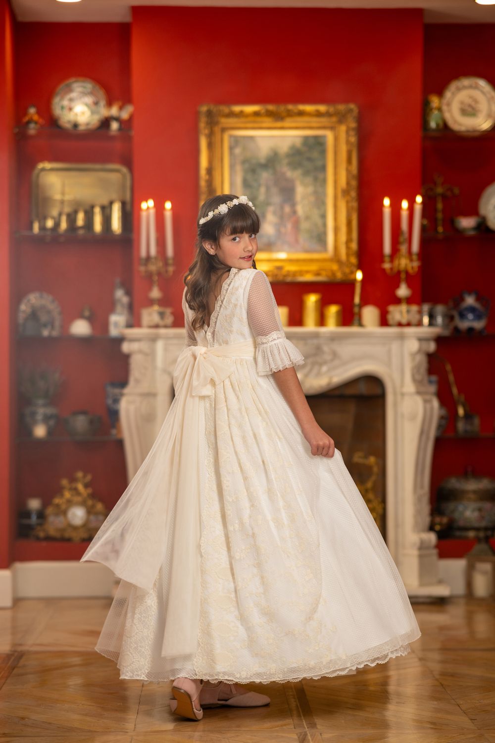 A young child in a flowing white christening gown spins gracefully on a hardwood floor in an elegant red room.