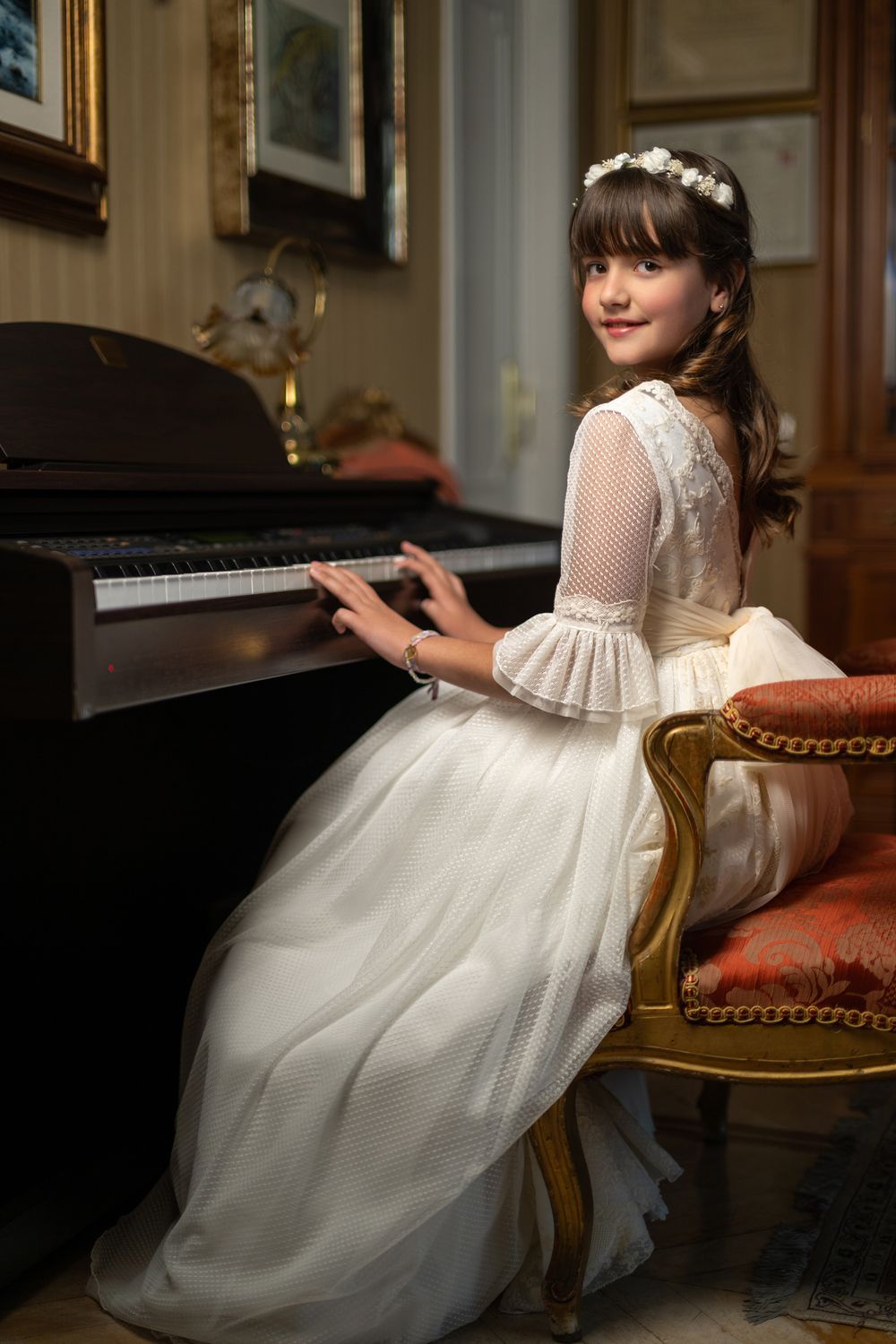 A young child in a white communion or flower girl dress sits gracefully on an antique chair in an elegant room.